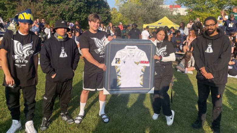 Five youth - four boys and one girl - pose around a framed jersey on a sunny day on a rugby field. They are all dressed in black sweat suits.