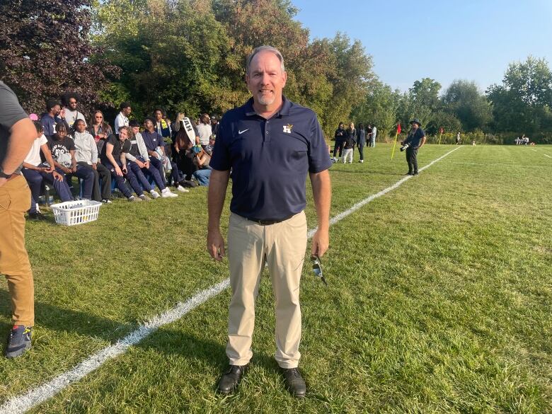 A middle-aged man with a goatee, in Humber College polo shirt, stands on a rugby field sideline on a sunny day. He is looking at the camera.