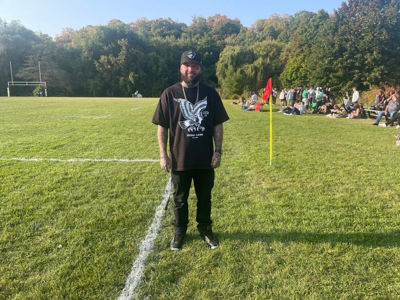 A young man in a ball cap and all black clothes stands on the sidelines of a rugby field on a sunny day. He is looking at the camera.