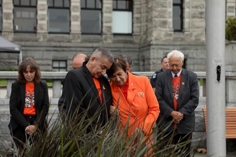 An Indigenous man and woman stand under a flag pole with their heads resting on each others shoulders. He wears a black suit with an orange shirt underneath and she wears a two-piece orange suit with an orange blouse underneath. 