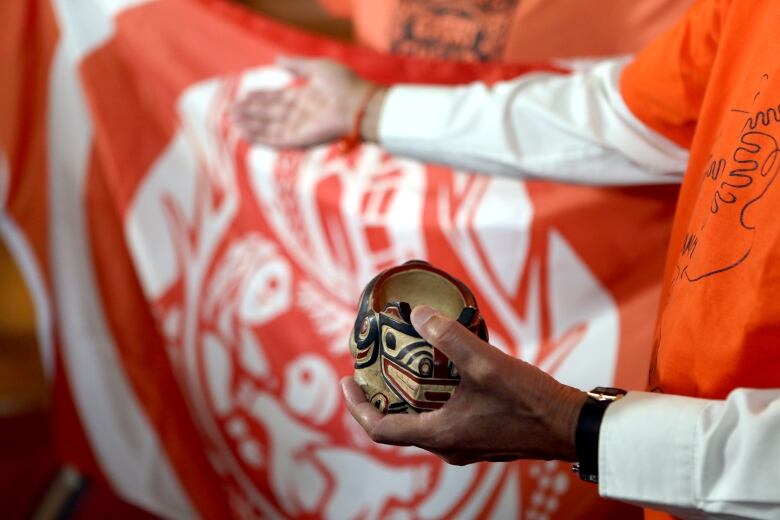 A wooden bowl painted with west coast Indigenous designs is shown in a man's hand in front of an orange and white flag.