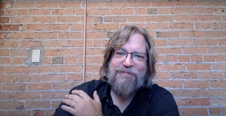 A man with light hair and a beard smiles at the camera in front of a brick wall.