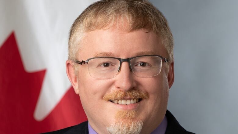 A man in glasses, suit and tie smiles in front of a Canadian flag.