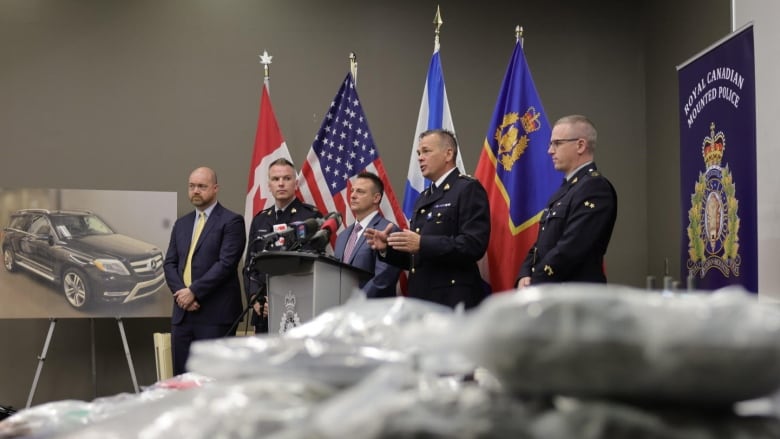 Bags are seen in the foreground, blurred, with five men in suits  standing at a podium in focus in the background.