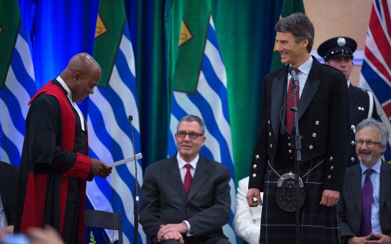 A Black man in judge's robes reads from a sheet of paper at a microphone while a white man stands smiling next to him wearing a kilt and black suit jacket with a red tie.