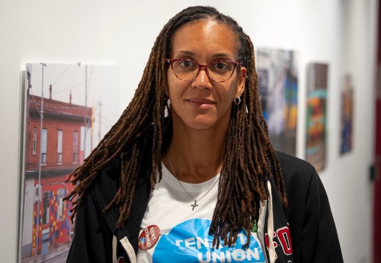 A woman with red glasses and long dark dreadlocks poses in a hallway with art hanging on the walls. She's wearing a t-shirt that says, 'tenant union' and a button that says, 'rent strike.'