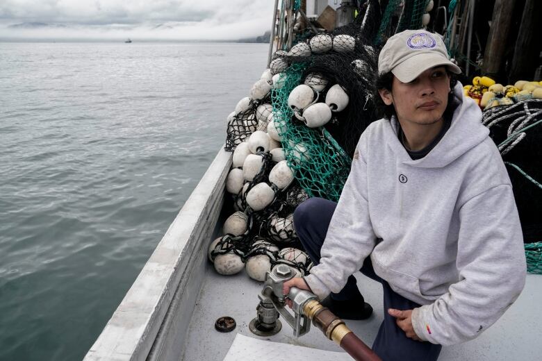 A man squats on the deck of a boat, fueling up, beside some large fishing nets.