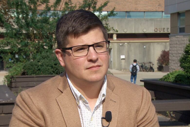 A man sits outside wearing a beige blazer and white button-up blouse.