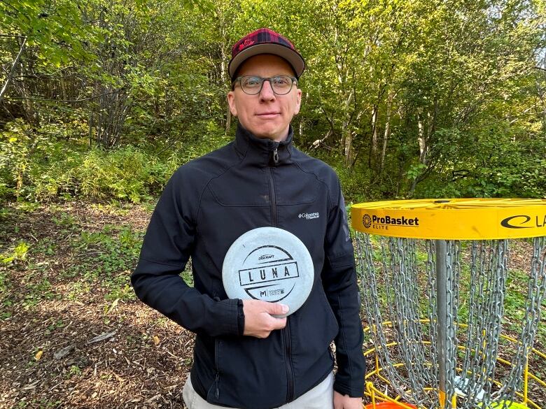 A man wearing outdoor wear holds a Frisbee in front of a large disc golf basket.