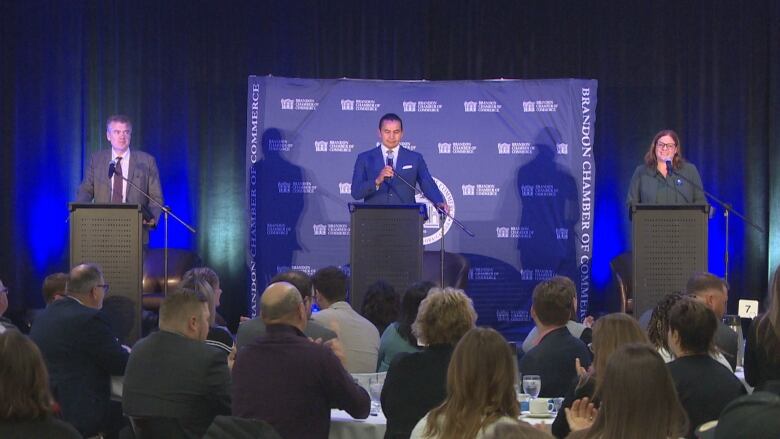 Three people stand at a podium, while people are seated in tables down below.