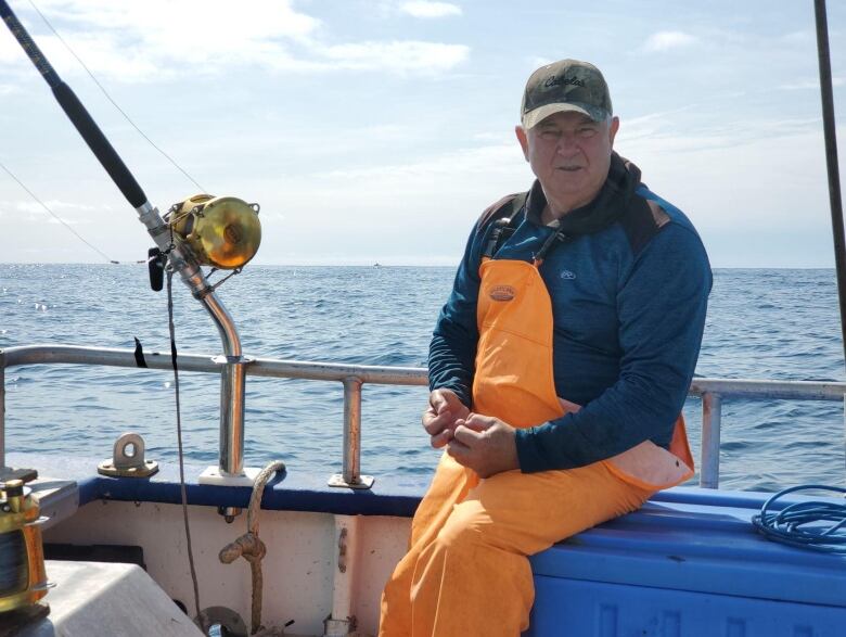 A man in a grey baseball cap, blue shirt and orange overalls sits on a boat in the water.