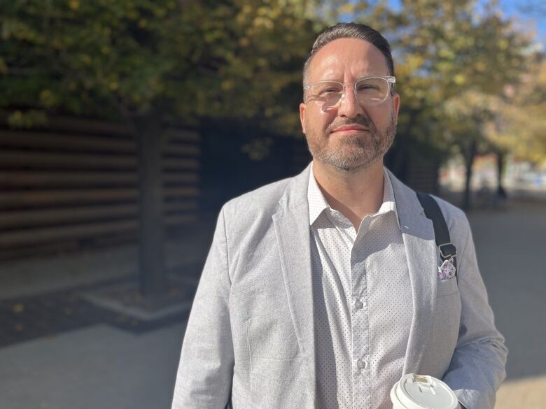 Man wearing clear glasses, light grey suit jacket and white shirt stands outside holding a coffee. 