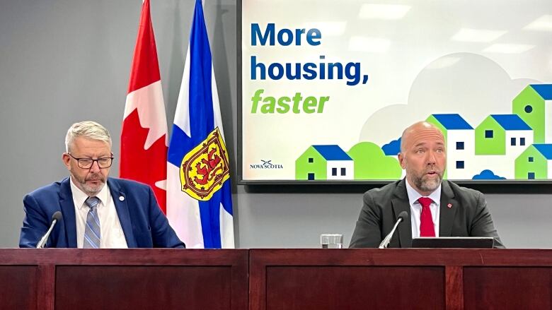 Two men sit at a table, with a Nova Scotia flag and Canadian flag in the background, as well as a poster that reads: More housing, faster.