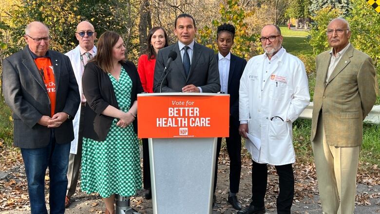 A man in a suit speaks at a podium with an orange sign that says 