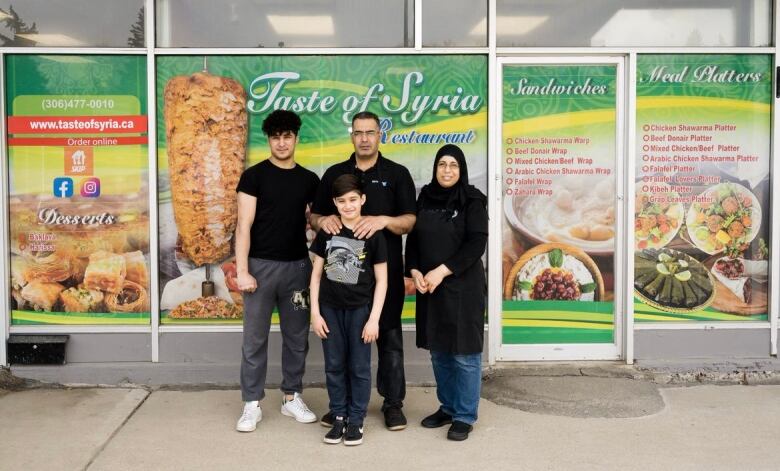 Two parents and two boys of different ages stand in front of a restaurant. Its windows are covered with decorative wraps that say 'Taste of Syria' and feature a variety of dishes.