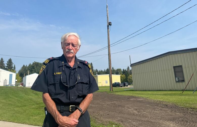 A gentleman with white hair and wearing a firefighter's dress uniform stands outside in front of a low building clad in aluminum siding.