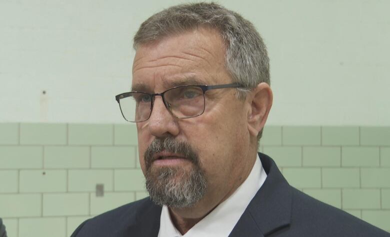 An unsmiling man with a grey beard and black-framed glasses