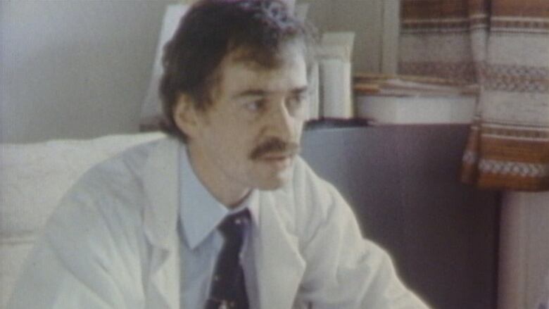 A man with a moustache and a white coat sits at a desk in an office.