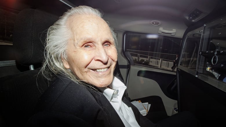 A man in his 80s, with long, swept-back white hair, wearing a black suit and white tie, smiles as he's shown in the back of a vehicle.