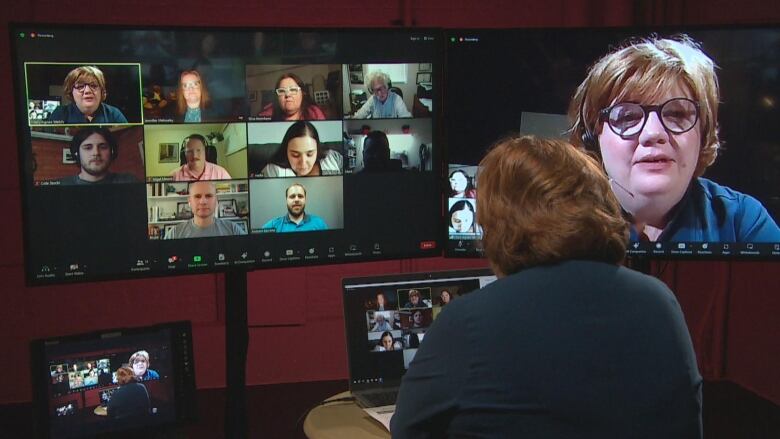 A woman is pictured from behind. In front of her are two screens, one shows a group of people on a Zoom call, and the other shows her face.