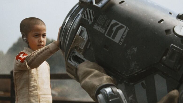 A young girl wearing a futuristic jump suit lays her hand against a robot bending over toward her. 