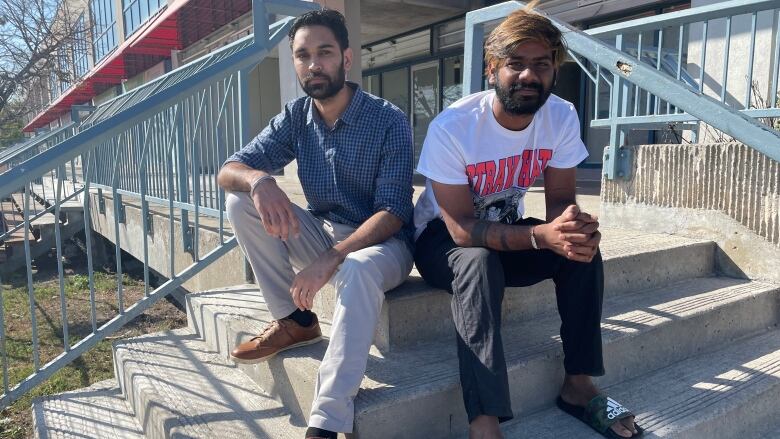 Two men sit on the steps of a building on a sunny day.