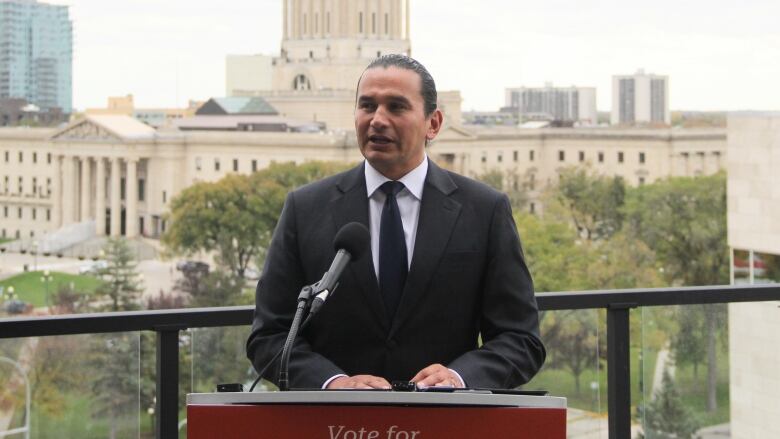 A man in a suit and tie stands on a balcony, overlooking the Manitoba Legislative Building in the background.