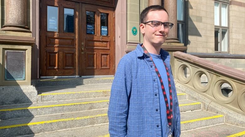 A white man with glasses and a blue plaid button up over a blue shirt looks at the camera. He stands in front of a stone building, with the door visible