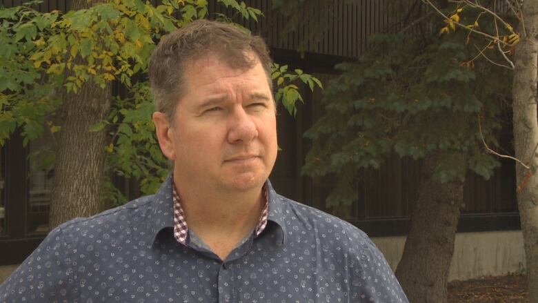 A man in a blue shirt is being interviewed, while a few trees are seen in the background.