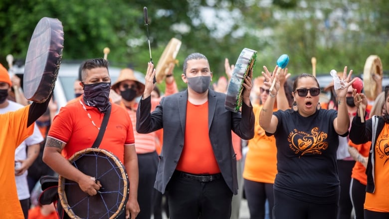 People in orange shirts stand holding drums, some wearing masks. 