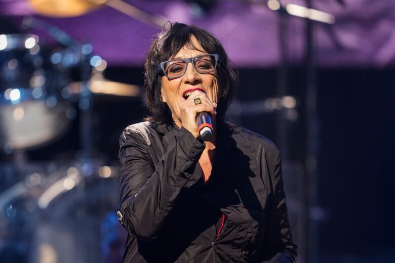 A woman wearing a black jacket and black-frame glasses sings on a stage.