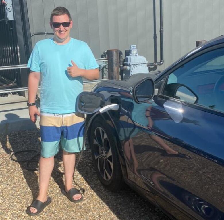 Man in a t-shirt and sunglasses gives a thumbs up beside a car charging at an EV charger