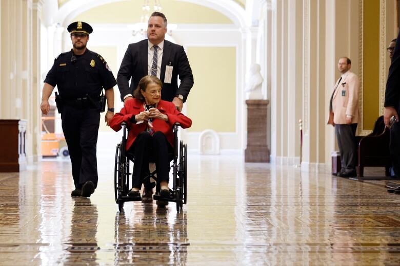 Woman in wheelchair being wheeled down a hall by a man who is accompanied by security. 