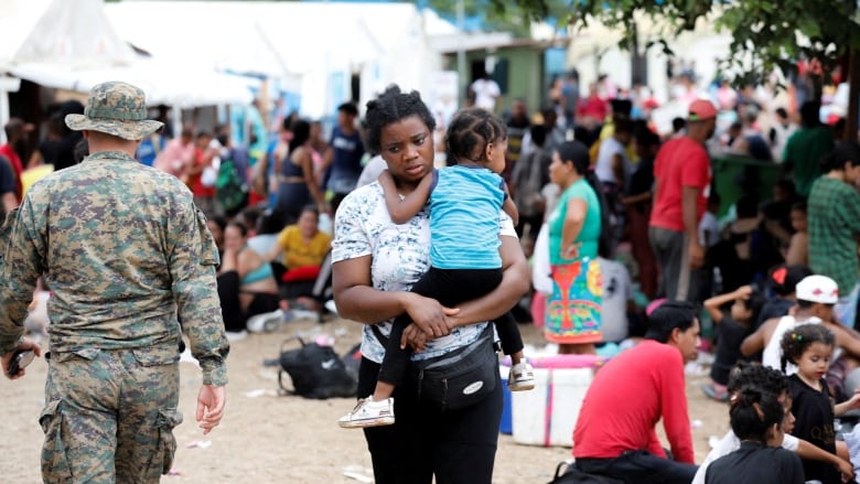 A woman holds a small child in her arms while standing outside, with tents and dozens of other people in the background.