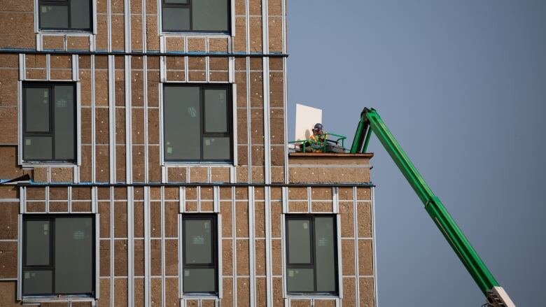 A construction worker on a green bucket lift installs equipment onto a building under construction.