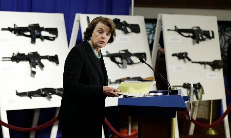 A woman is shown speaking before a display of bulletin boards depicting assault weapons.