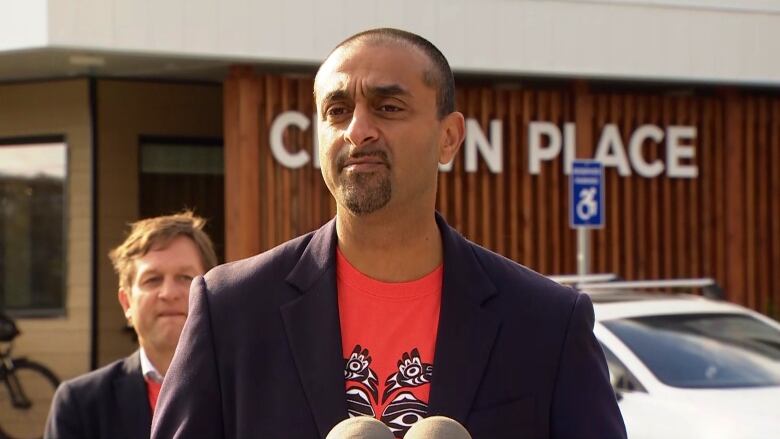 B.C. Housing Minister Ravi Kahlon wears a suit jacket with a orange shirt underneath and speaks into a microphone. B.C. Transportation Minister Rob Fleming stands in the background.