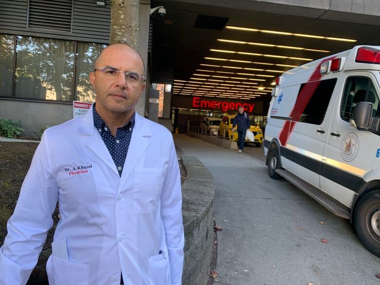 A bald man wears glasses and a white doctor's robe stands outside an emergency department.