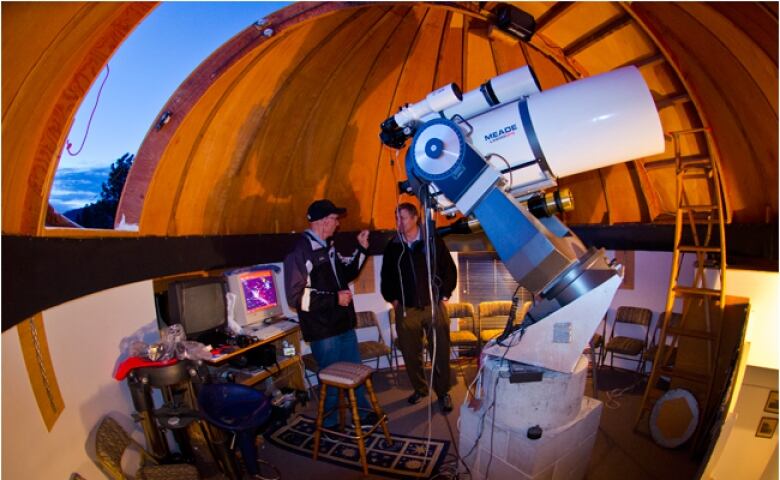 Two men stand in a circular observatory room beside a large round telescope.