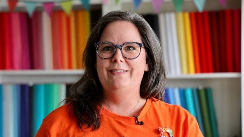 A woman wearing an orange T-shirt and glasses smiles. There are rows of colourful fabric in the background.