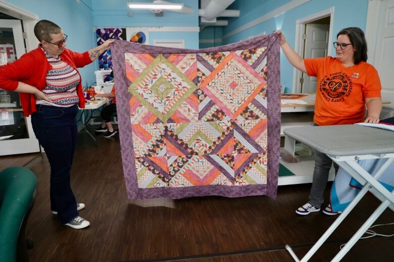 Two people hold up a large, colourful quilt top in a quilting studio.