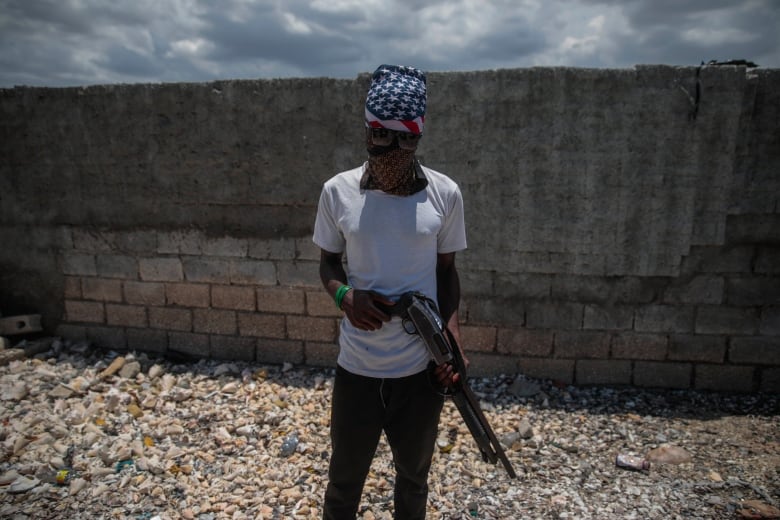 A gang member wearing sunglasses and a face covering holds a shotgun.