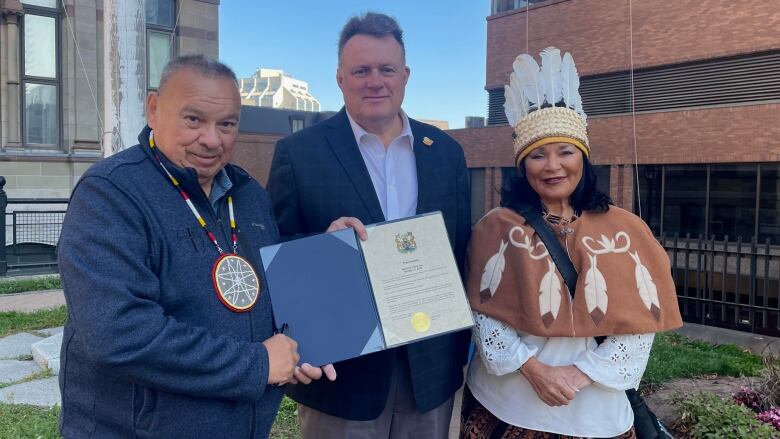 Three people pose for a photo holding an official document. Two men on the left, and a woman on the right. The woman wears Indigenous regalia.