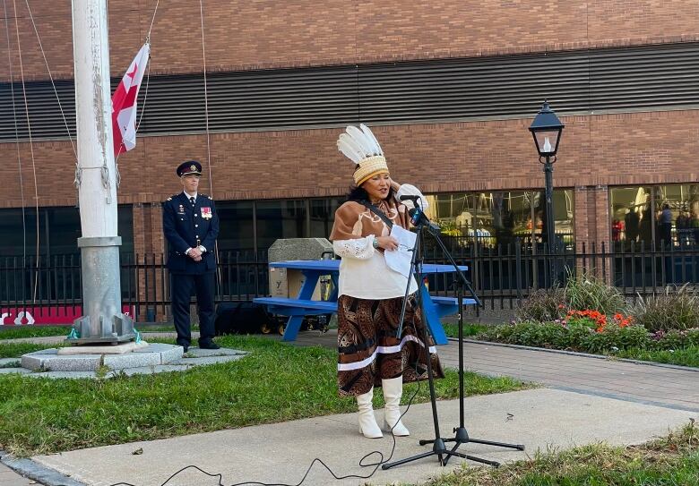 A woman gives a speech at a microphone stand. She is wearing brown and white Indigenous regalia.