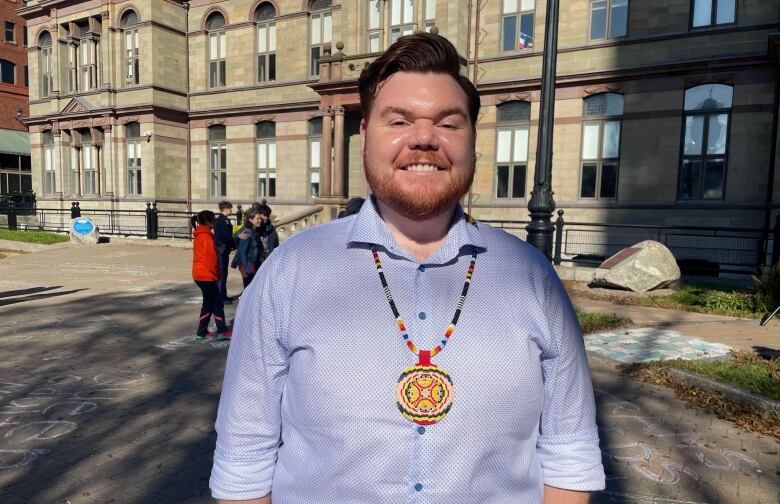 A man is smiling at a camera. He wears a blue button-up shirt. He has reddish hair and a beard. He is wearing a beaded piece of Indigenous regalia around his neck.