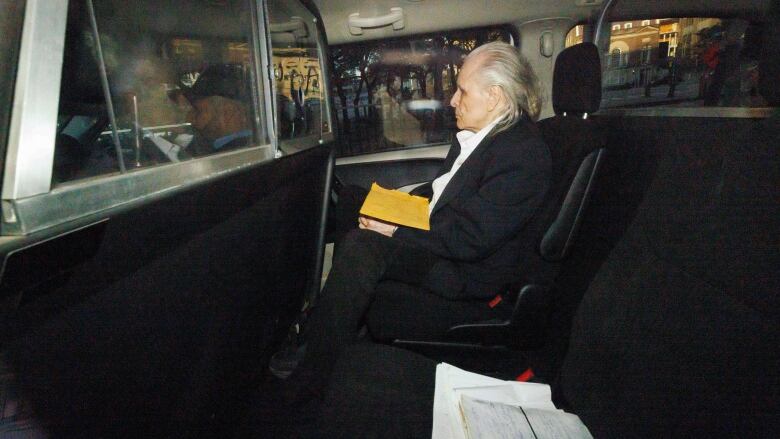 An older man with white hair, wearing a black suit and white shirt, sits with his legs crossed in the back of a vehicle.