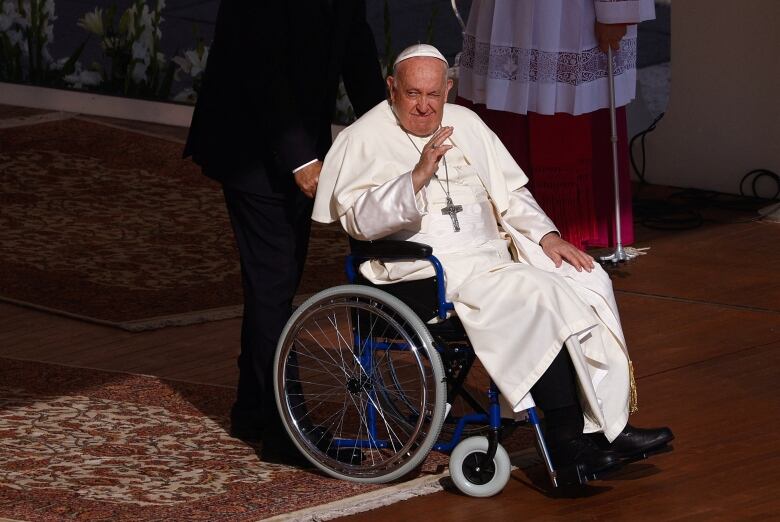 The Pope is shown in a wheelchair wearing white papal garments and cap.