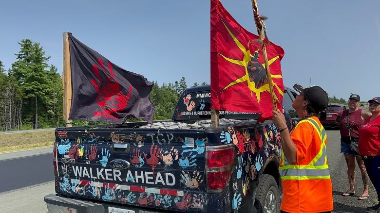 A person wearing an orange safety vest stands in front of a truck covered in painted handprints. Two flags stick up out of the truck, which is parked on the side of the road.