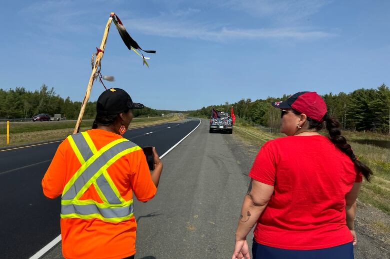 Two women walk side-by-side down the side of a highway, a pick-up truck in the distance. The woman on the left wears an orange safety vest and holds up a flag. The woman on the right wears a red t-shirt and baseball cap.