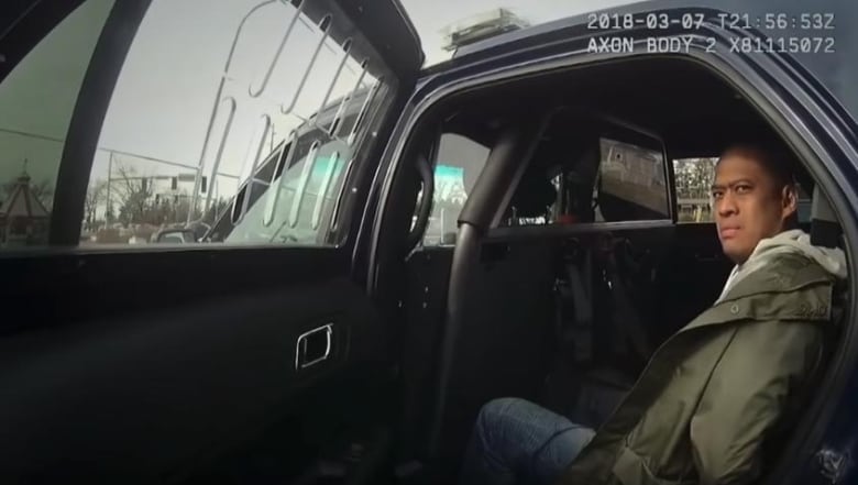 A bald man sitting in a police cruiser with the rear passenger door open.